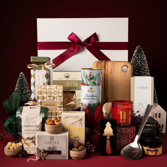 white gift box on display next to wooden board, red candle, assorted nuts, silver salad servers and christmas tree decorations