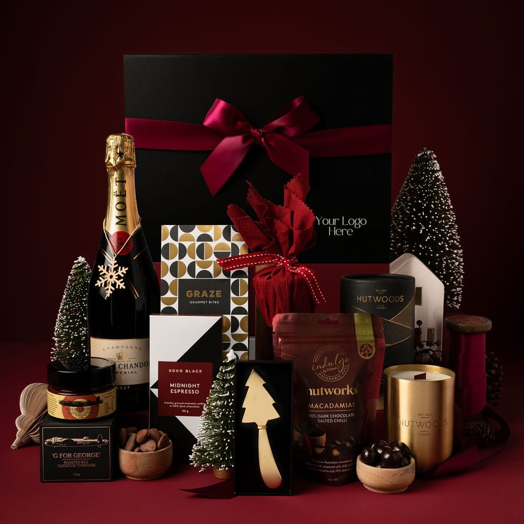 black gift box and red ribbon on display next to champagne, gold candle, cheese knife, chocolate and pudding with christmas tree decorations and red back drop