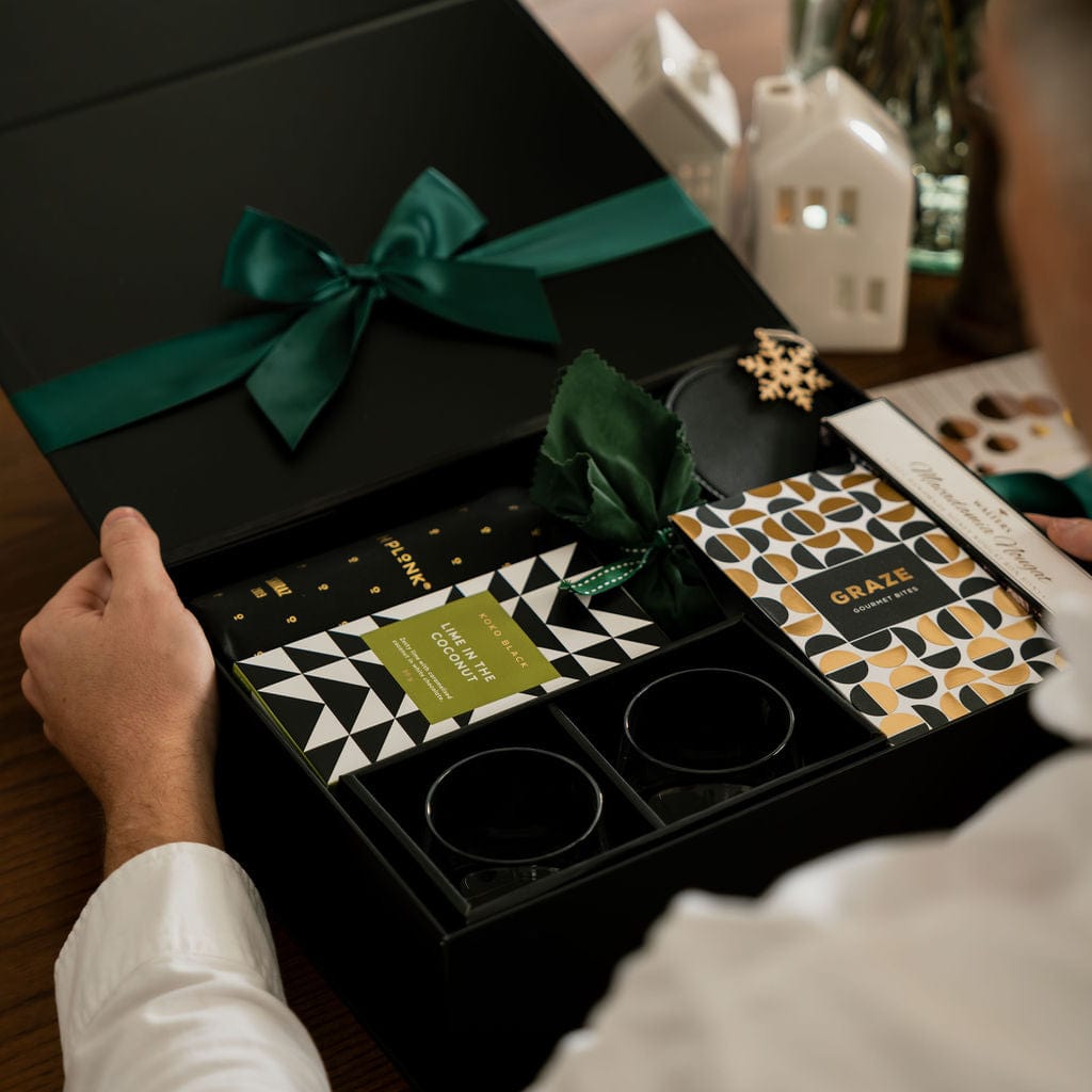 man holding open gift in black box showing glasses, chocolate and green ribbon