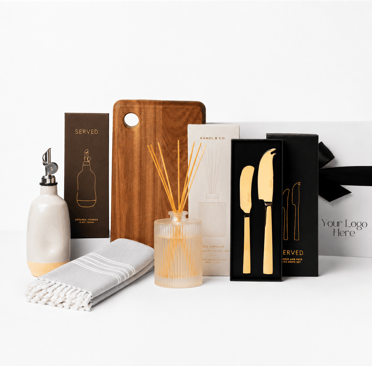 A curated gift set on a white background featuring a white ceramic oil bottle with a silver pourer, a wooden cutting board, a reed diffuser with a frosted glass bottle and reeds, a folded white fringed cloth, a boxed knife set with gold-toned handles, and branded packaging.