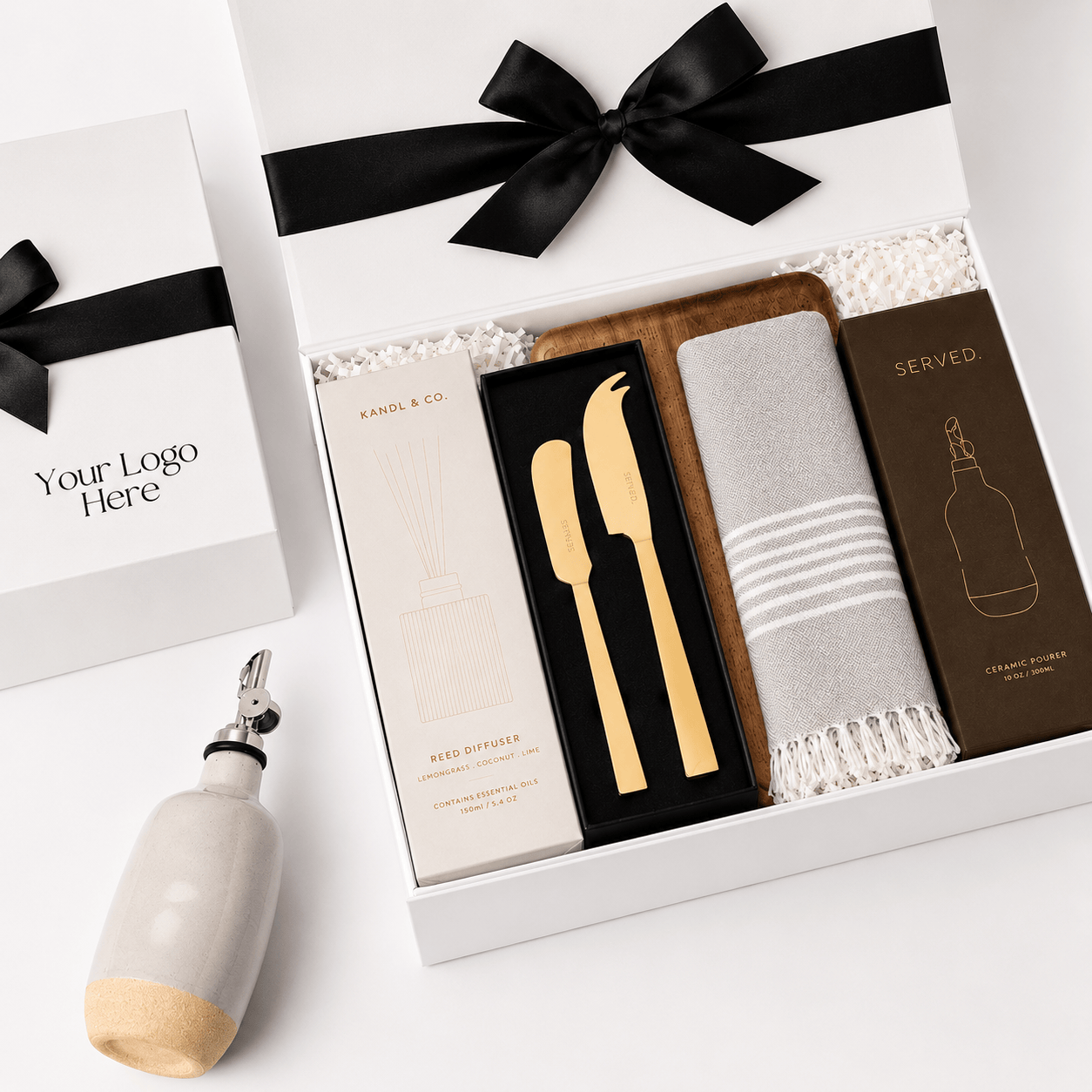 Elegant Abode open white gift box with a black ribbon and logo placeholder, containing a Kandl & Co reed diffuser, a SERVED gold cheese and pate knife set, a grey striped Turkish hand towel and a ceramic oil pourer on a white background