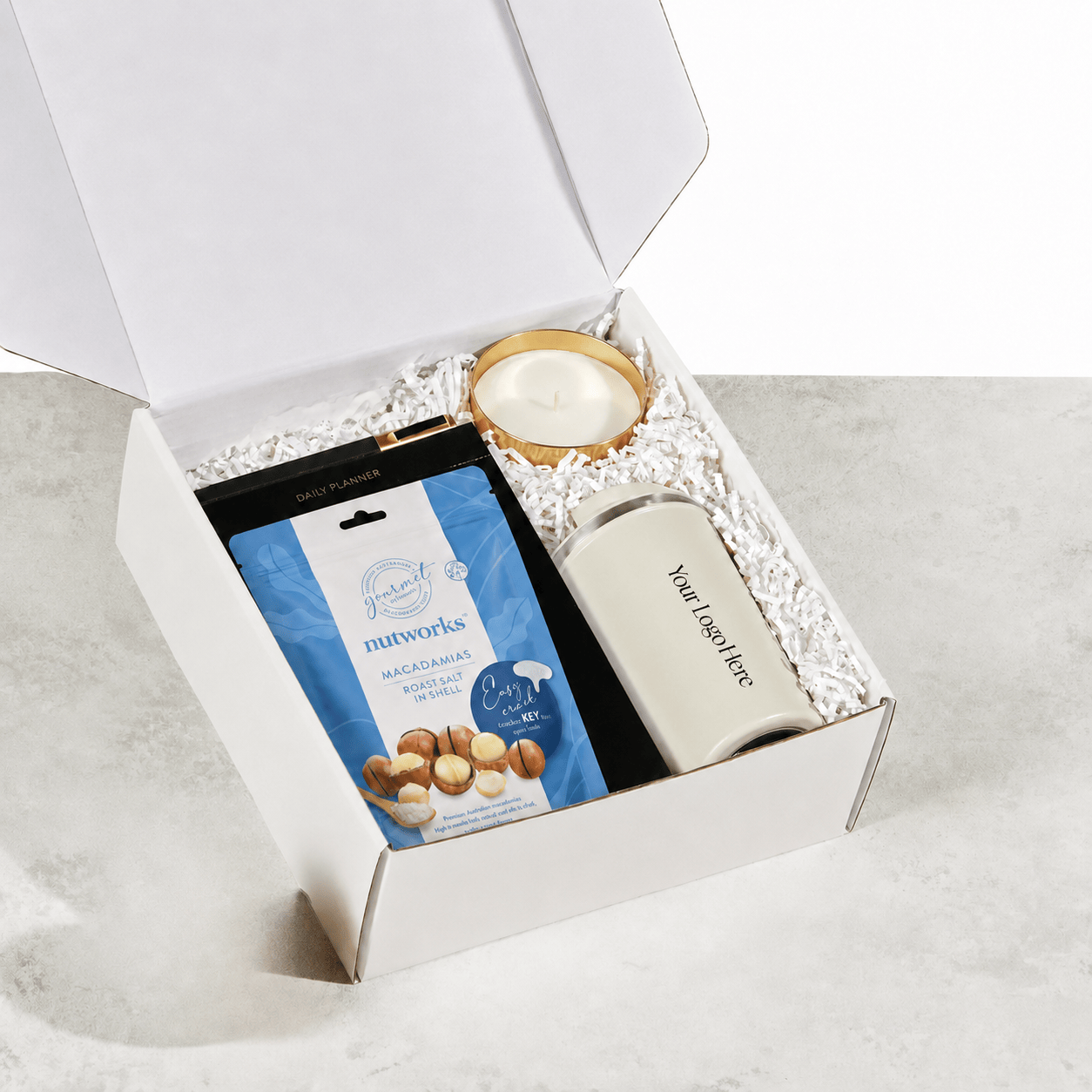 An open white gift box on a light stone surface, filled with white crinkle paper and containing a blue bag of Nutworks Calamansi Almond Snacks, a cream-colored cylindrical bottle with placeholder text, and a round gold-rimmed candle.