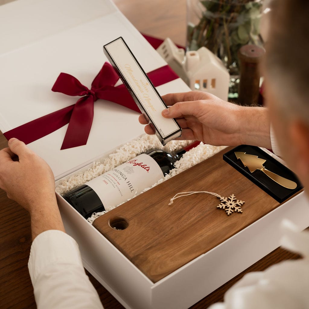 man holding nougat next to open gift with red ribbon, wine, snow flake ornament and chopping board