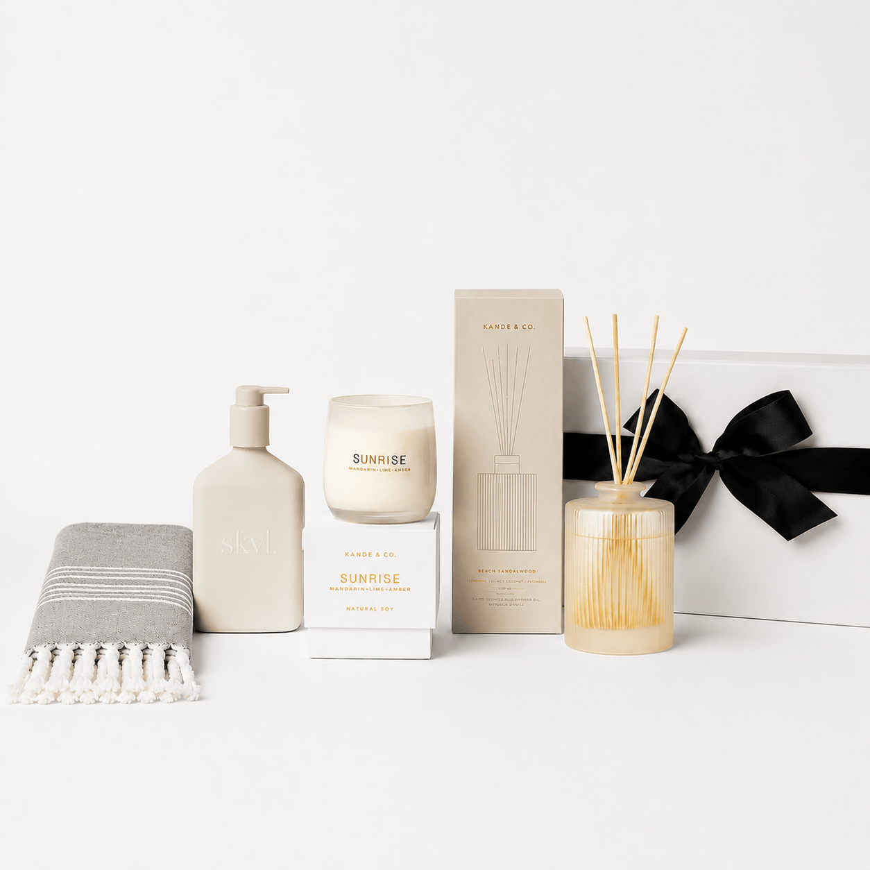 A curated gift set featuring a beige skincare pump bottle, a candle in a glass jar, a reed diffuser with wooden reeds, a folded gray throw blanket with white fringe, and a white gift box tied with a black ribbon. The items are arranged on a clean white surface against a white background.