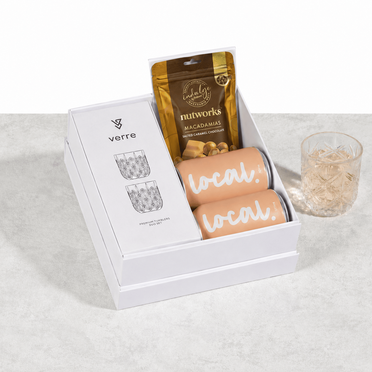 A gift set in a white box featuring two patterned glass tumblers, a pouch of Nutworks Macadamia Flavored Energy Gummies, and two peach-colored canes or sticks with the word 'local.' printed on them, alongside a clear glass of iced coffee on a light gray surface.