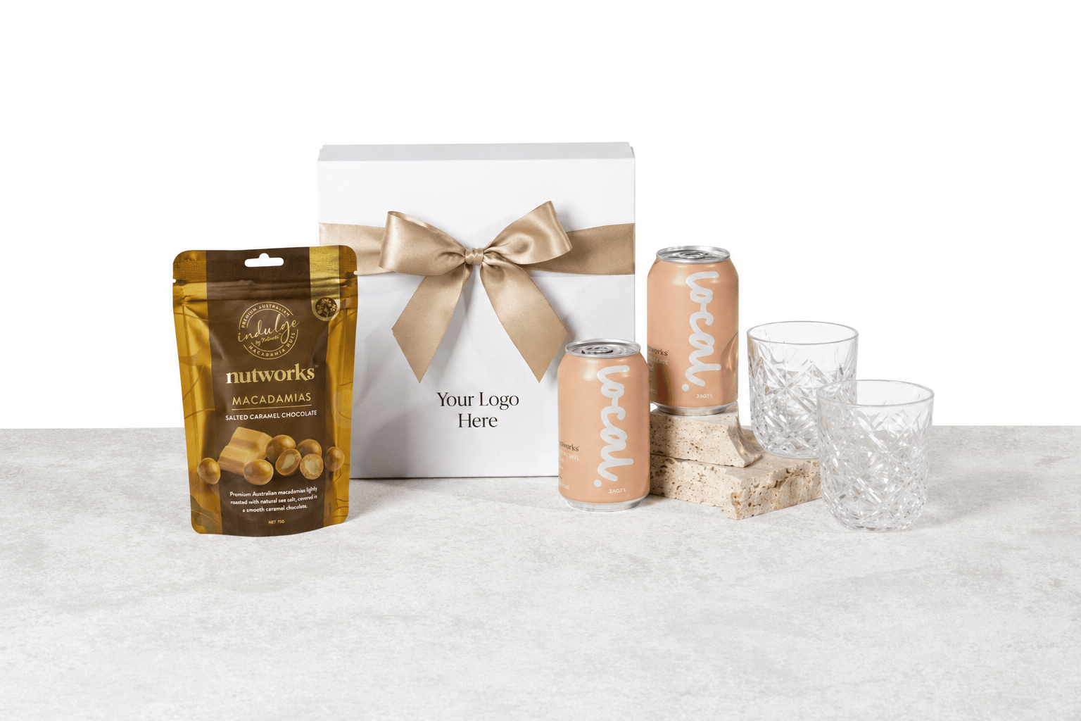 Sip & Savour gift box with a beige ribbon and logo placeholder, shown with a pouch of Nutworks salted caramel macadamias, two LOCAL peach vodka iced tea seltzer cans and two VERRE crystal tumbler glasses on a light grey surface
