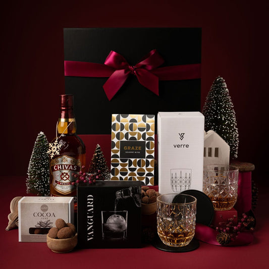 christmas gift box with whiskey, glasses chocolate and ice moulds on display in front of red backdrop 