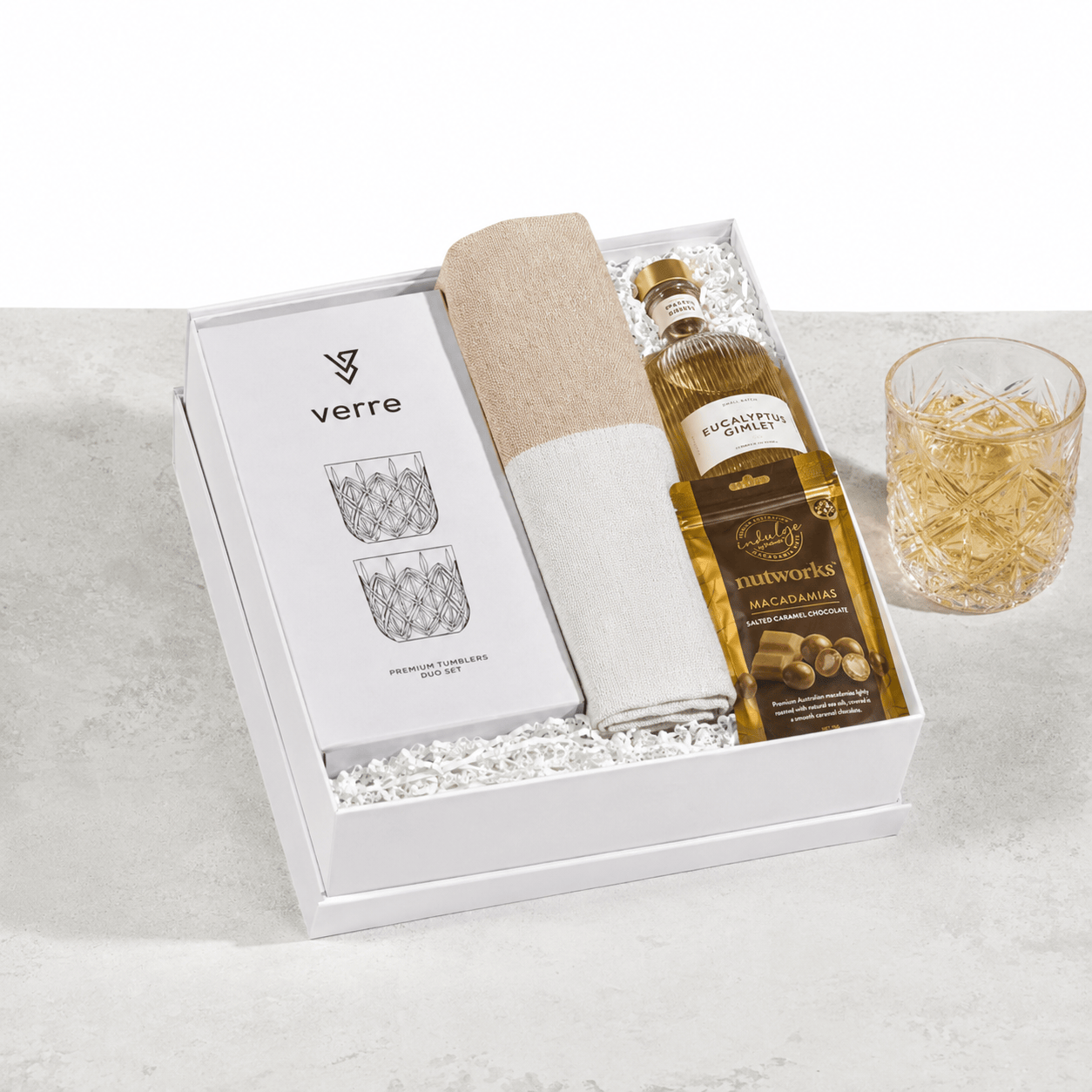 The Peaceful Pour open gift box containing a VERRE premium tumbler set, a rolled cream Turkish hand towel, a MAYBE SAMMY Eucalyptus Gimlet bottle and a Nutworks salted caramel macadamias pouch, with a filled crystal tumbler alongside on a light grey surface