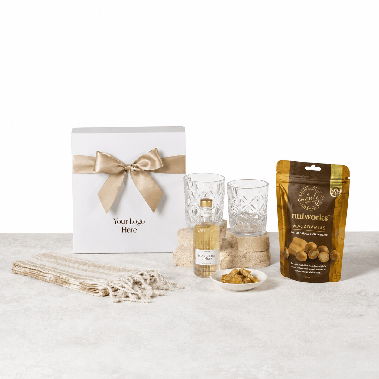 The Peaceful Pour gift box with a beige ribbon and logo placeholder, shown with a MAYBE SAMMY Eucalyptus Gimlet bottle, two VERRE crystal tumblers, a Nutworks salted caramel macadamias pouch and a cream Turkish hand towel on a light stone surface