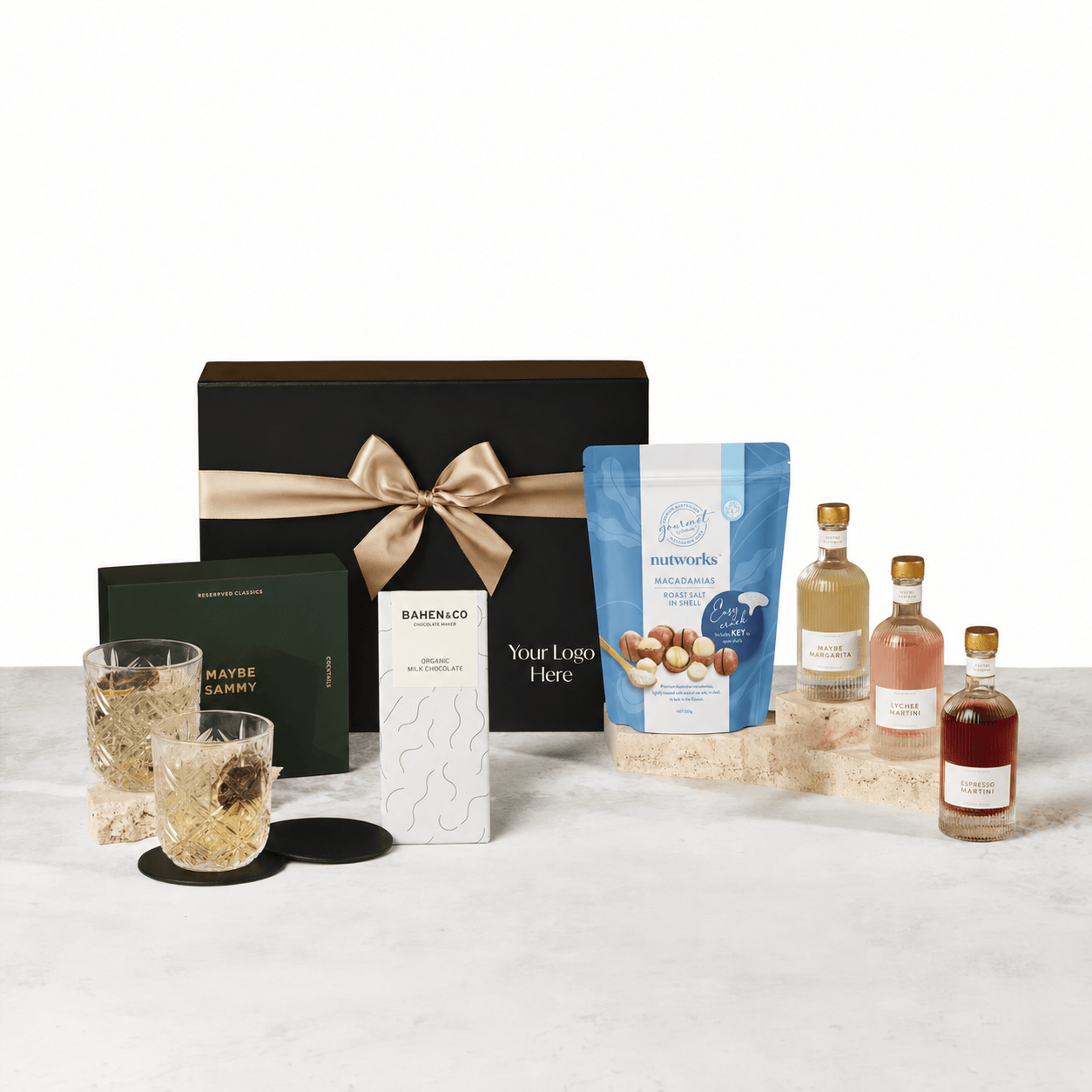 The Tasty Tipple black gift box with a beige ribbon and logo placeholder, shown with a MAYBE SAMMY cocktails trio pack, two VERRE crystal tumblers, round leather-look coasters, Bahen & Co chocolate and a Nutworks macadamias pouch on a marble surface