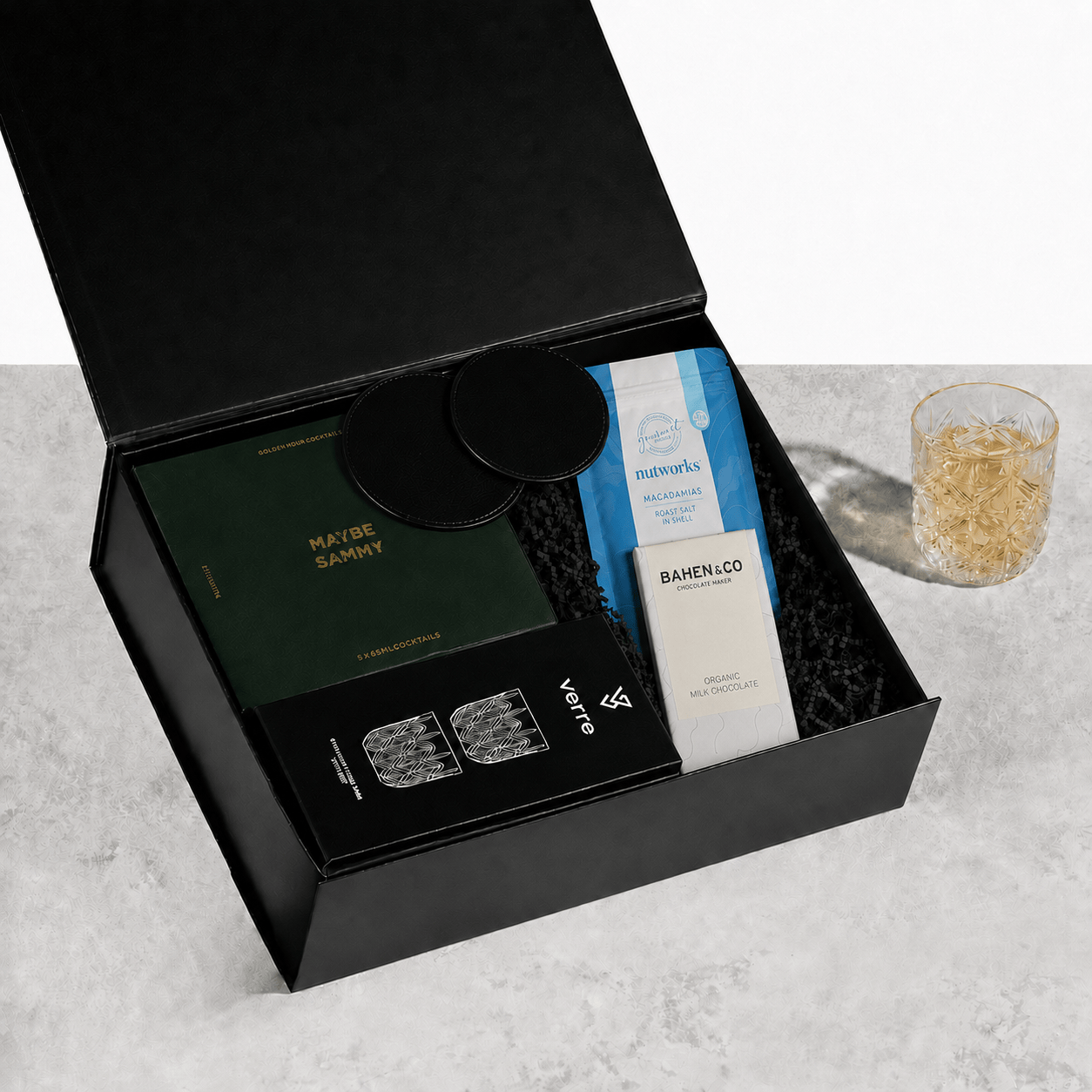 The Tasty Tipple open black gift box containing a MAYBE SAMMY cocktails box, round leather-look coasters, a Nutworks macadamias pouch, Bahen & Co chocolate and a VERRE tumbler set, with a filled crystal tumbler alongside on a light stone surface
