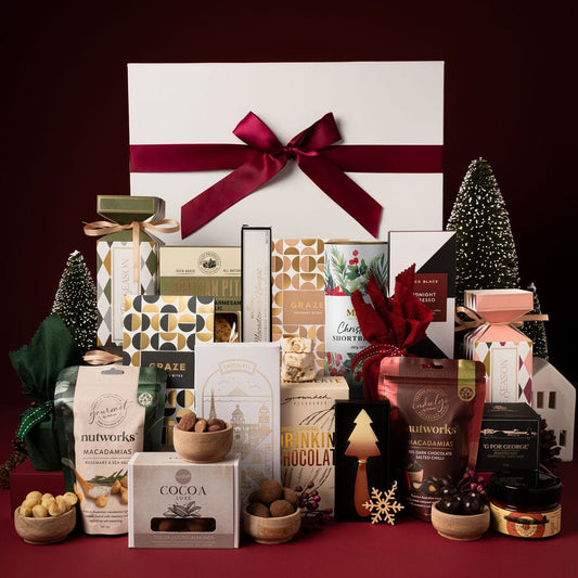 white gift box with red ribbon and contents on display including chocolate, nuts, christmas bon bon, gold cheese knife and shortbread against red backdrop