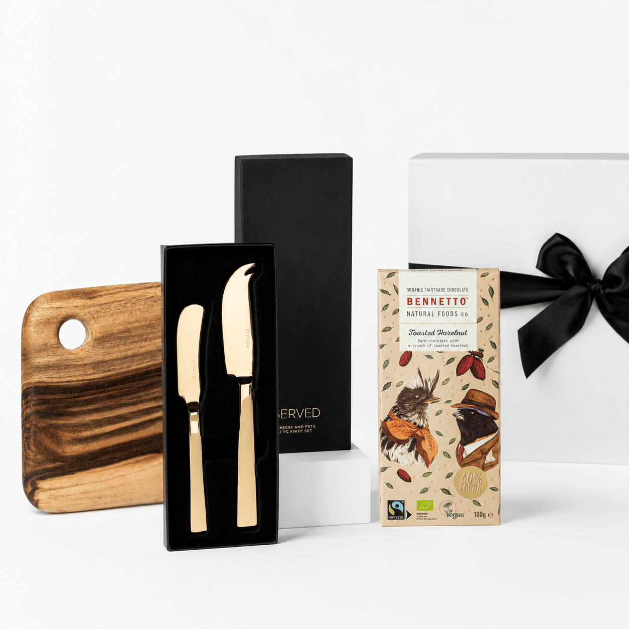 Well Wishes gift set with a white box and black ribbon, shown with a Camphor Laurel wooden serving board, a SERVED gold cheese and pate knife set and a Bennetto Natural Foods toasted hazelnut chocolate bar on a white background