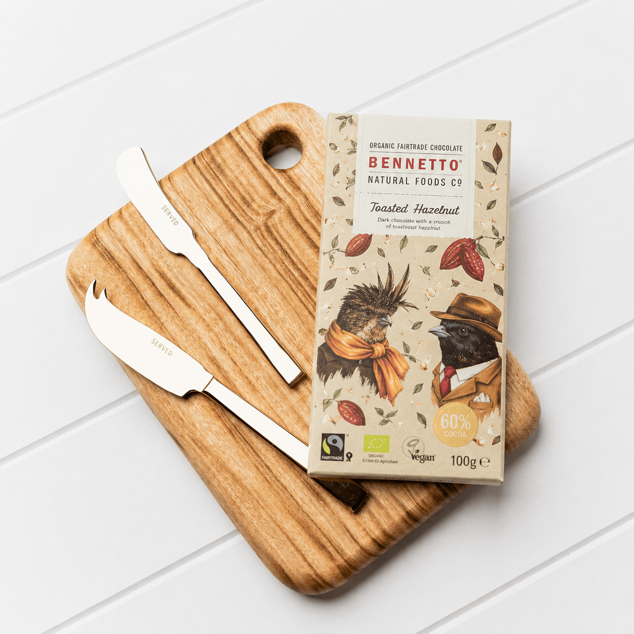 Bennetto Natural Foods toasted hazelnut chocolate bar and a SERVED gold cheese and pate knife set on a Camphor Laurel wooden serving board on a white panelled surface
