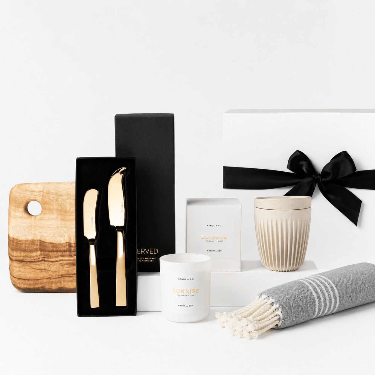 Welcome Home gift set with a white box and black ribbon, shown with a Camphor Laurel wooden serving board, a SERVED gold cheese and pate knife set, a Kandl & Co candle, a ribbed reusable cup and a grey striped Turkish hand towel on a white background