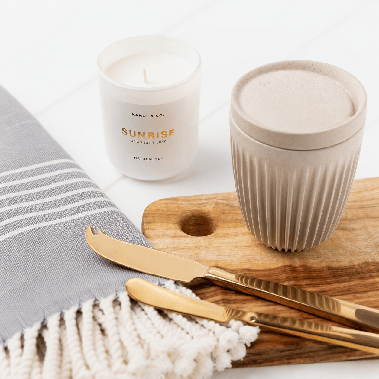 Styled close-up of a Kandl & Co Sunrise coconut and lime candle, a ribbed reusable cup and a SERVED gold cheese and pate knife set on a Camphor Laurel wooden board with a grey striped Turkish hand towel