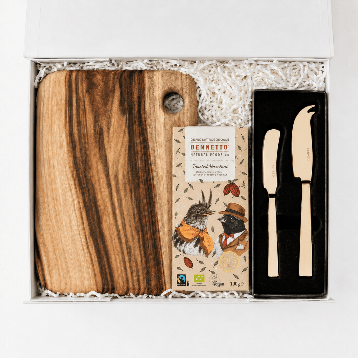 Well Wishes open white gift box containing a Camphor Laurel wooden serving board, a Bennetto Natural Foods toasted hazelnut chocolate bar and a SERVED gold cheese and pate knife set on white crinkle filler